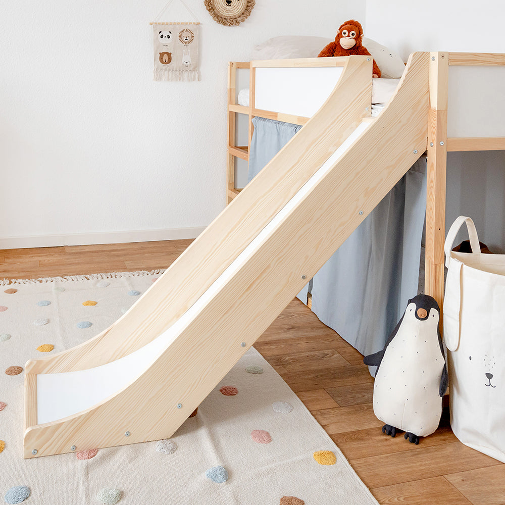 Seitliche Ansicht einer Rutsche fürs Ikea Kura Hochbett in einem dekorierten Kinderzimmer. Die Rutsche und das Bett sind aus Kiefernholz hergestellt. Zudem sind am Kura Bett zusätzlich blaue Vorhänge angebracht.