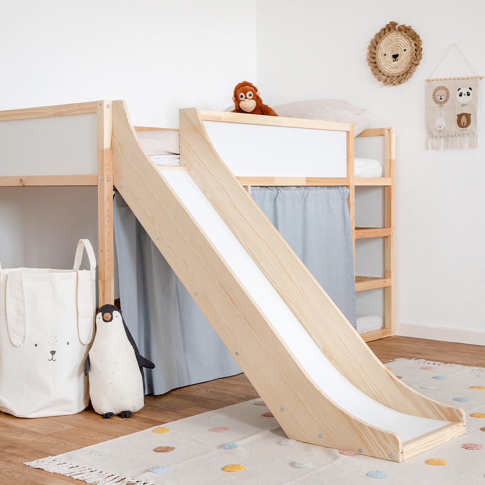 Seitliche Ansicht einer Rutsche fürs Ikea Kura Hochbett in einem dekorierten Kinderzimmer. Die Rutsche und das Bett sind aus Kiefernholz hergestellt. Zudem sind am Kura Bett zusätzlich blaue Vorhänge angebracht.