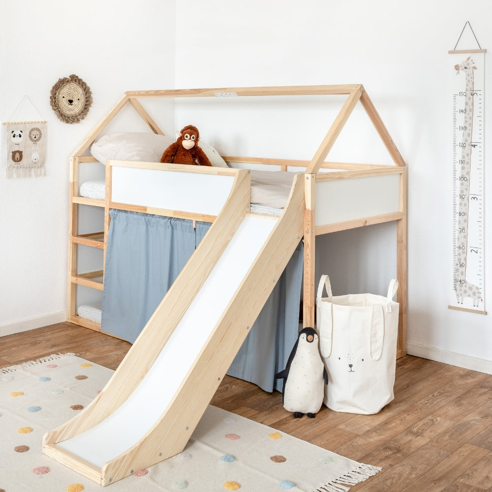 Kura Bett von Ikea als Hochbett im dekorierten Kinderzimmer mit blauen Vorhängen, einem Dachgestell aus Holz und einer passgenauen Rutsche. Durch das Zubehör wirkt das Kura Bett wie ein Hausbett und die Kinder können sich in der unteren Ebene hinter den Vorhängen verstecken. 