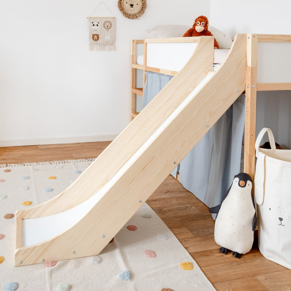 Seitliche Ansicht einer Rutsche fürs Ikea Kura Hochbett in einem dekorierten Kinderzimmer. Die Rutsche und das Bett sind aus Kiefernholz hergestellt. Zudem sind am Kura Bett zusätzlich blaue Vorhänge angebracht. 